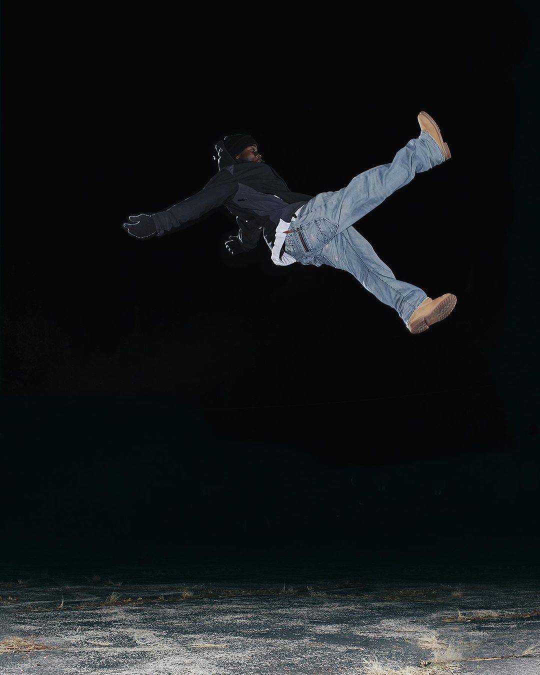 Man in mid-air wearing jeans, jacket, and beanie against a dark background with a concrete-like surface. Athletic pose in nighttime setting.