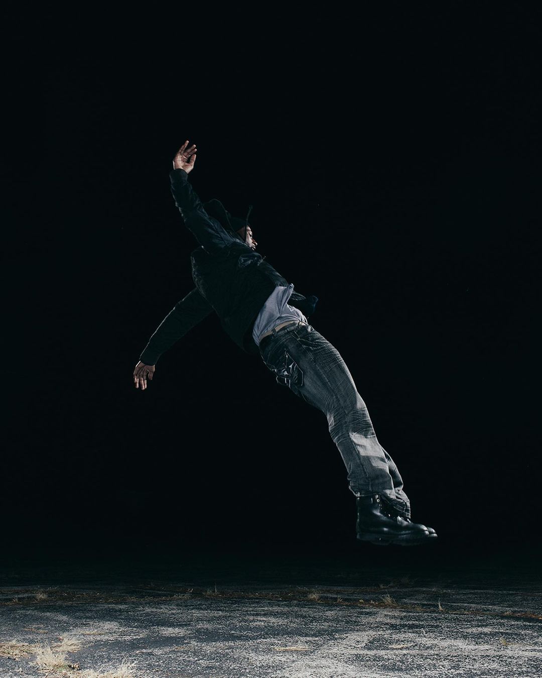 Man wearing jeans and a jacket falls through the air against a dark background. He is suspended above a textured grey surface.