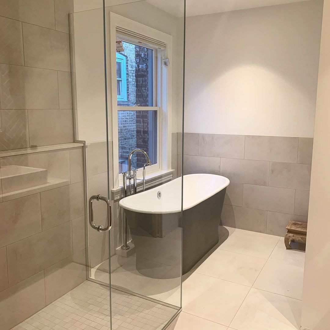 Freestanding bathtub in a minimalist bathroom with beige tile, glass shower, and a view from the window, bathed in natural light.