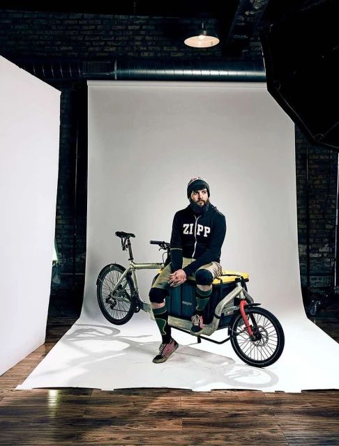 Bearded man in ZIPP hoodie sits on a cargo bike in photo studio with white backdrop, brick wall, wearing beanie, knee pads, patterned socks.