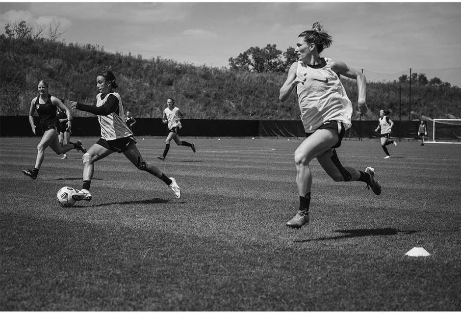 Black and white shot of women's soccer team during a training session on a sunny field. Players run, kick a soccer ball, trees and sky.