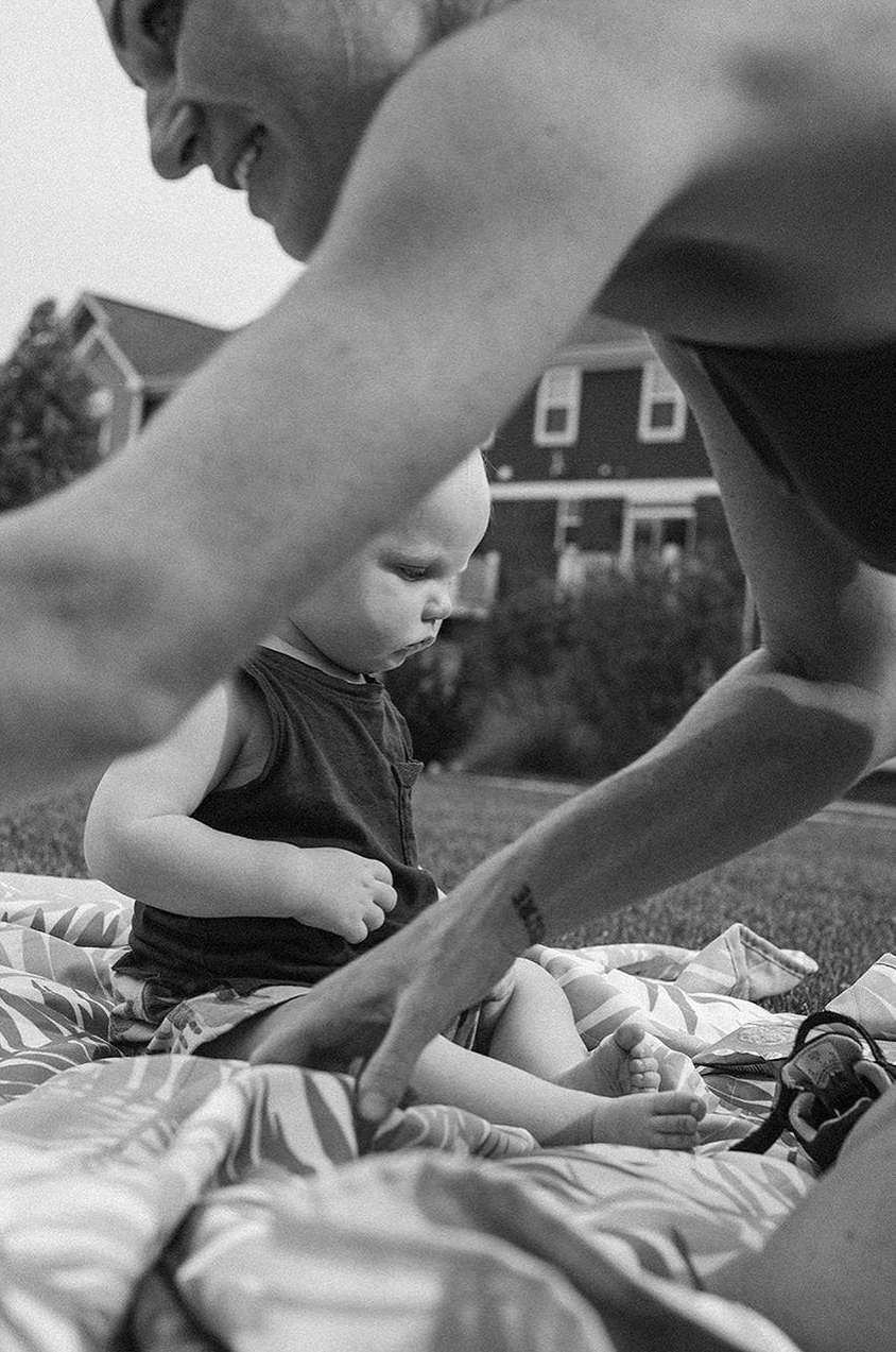 A close black and white photo of a baby sitting on a patterned blanket outdoors, with an adult's hands guiding the infant.
