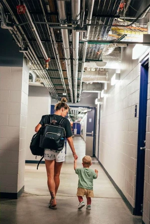 Woman and toddler walking away down an indoor hallway, woman carries a backpack and the child holds her hand.