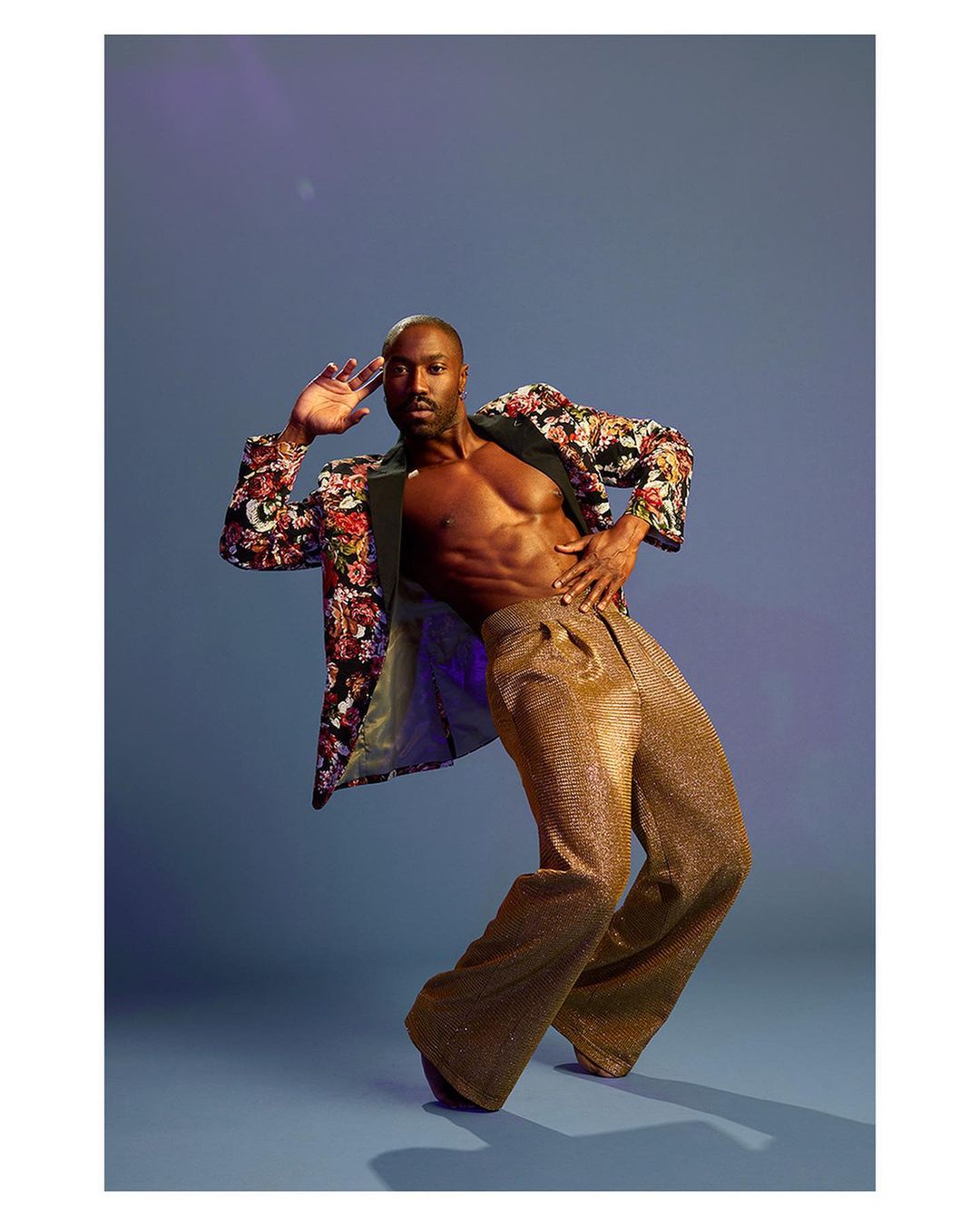 Portrait of an adult Black male in gold pants and a floral jacket in a dance pose against a blue background with purple lighting.