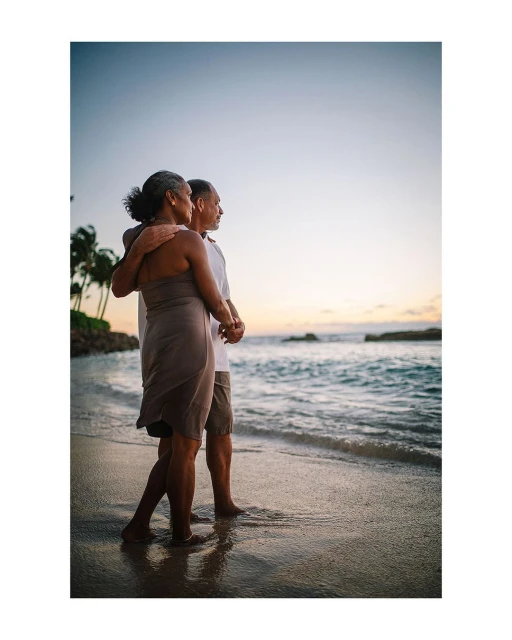 Elderly couple embracing at sunset on a tropical beach, enjoying the ocean view. Warm light highlights their figures and the tranquil scene.