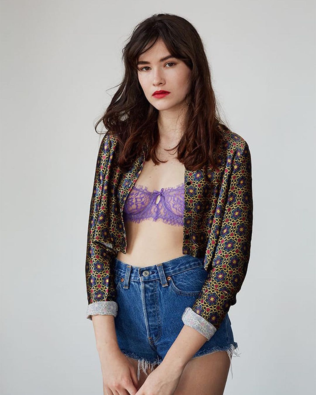 Model wearing patterned open jacket, purple lace bra and denim shorts against a plain white studio wall.