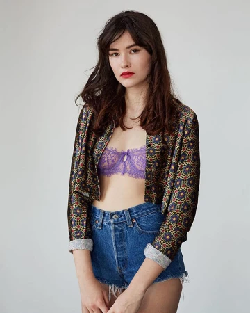 Model wearing patterned open jacket, purple lace bra and denim shorts against a plain white studio wall.