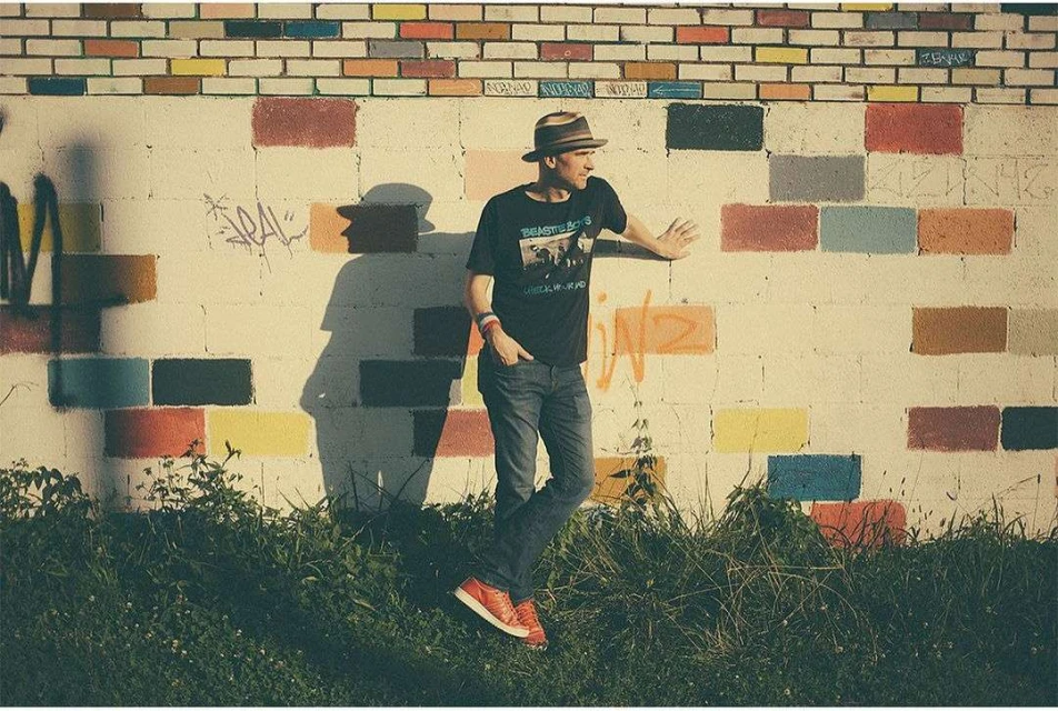 Adult male with hat standing in front of colourful brick wall wearing jeans, t-shirt and red shoes, shadowed by natural light.