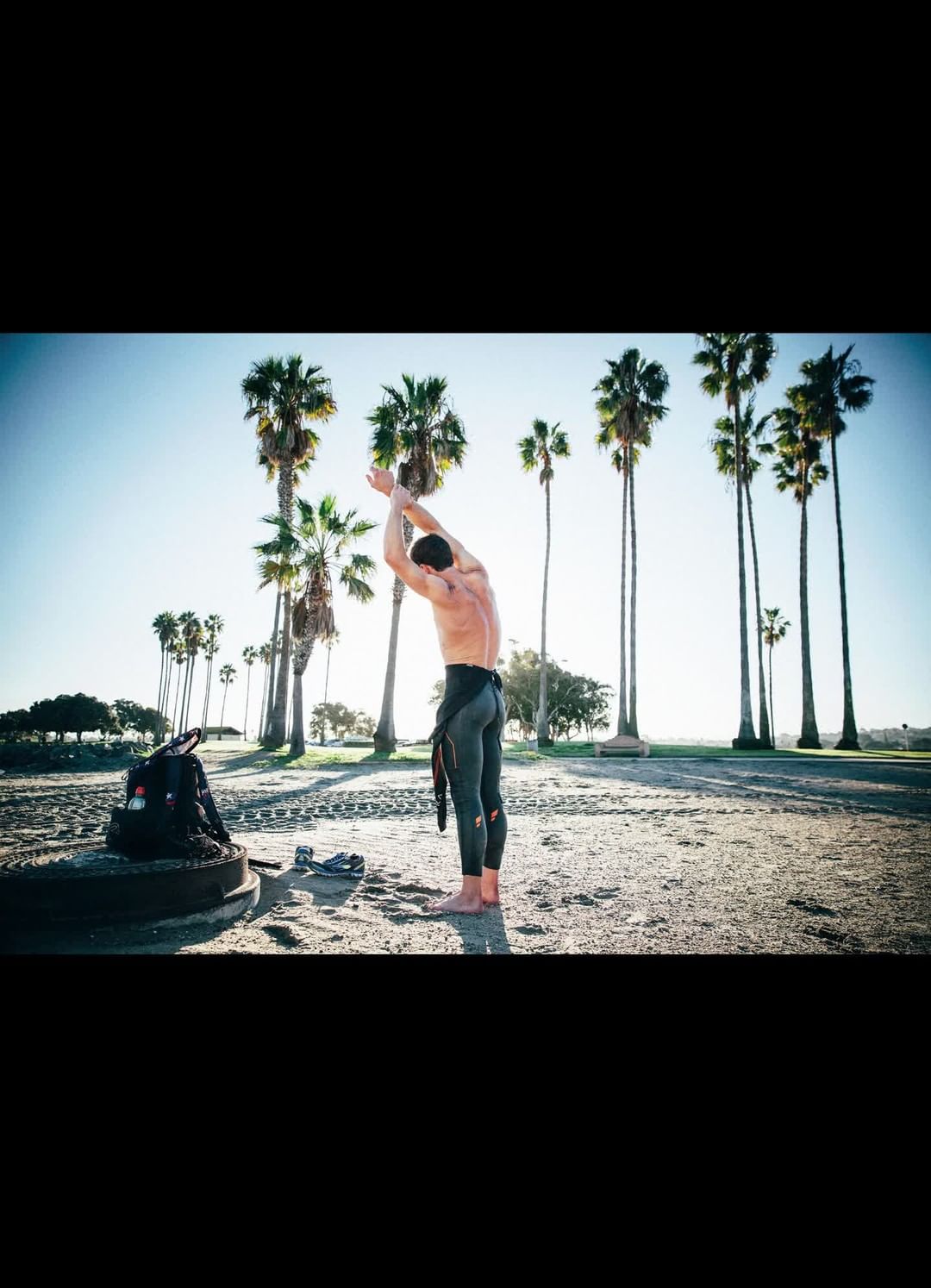 Man in wetsuit stretches on sandy beach near backpack and palm trees under bright sky, capturing a fitness moment in nature.
