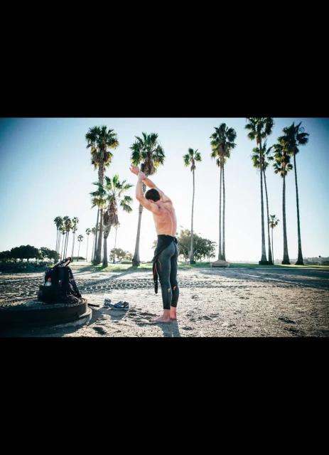 Man in wetsuit stretches on sandy beach near backpack and palm trees under bright sky, capturing a fitness moment in nature.
