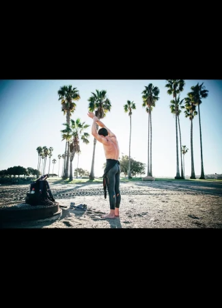 Man in wetsuit stretches on sandy beach near backpack and palm trees under bright sky, capturing a fitness moment in nature.