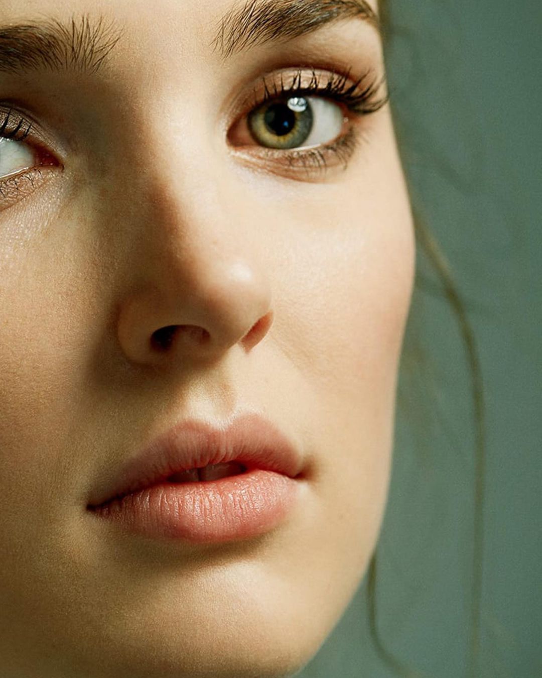 Close-up portrait of a fair-skinned woman with hazel eyes, focusing on her facial features against a muted teal background.