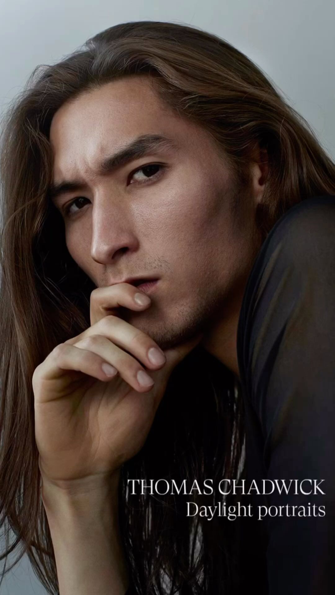 Close-up portrait of a man with long brown hair and dark shirt touching his chin against a plain light grey background.