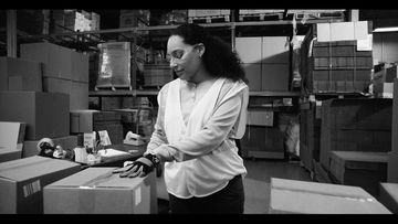 Monochrome image of a female warehouse worker in a safety vest checking a shipping box surrounded by stacks of boxes in a warehouse.