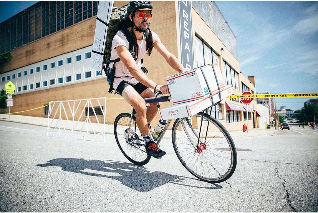Adult male on a bicycle with boxes attached to the front, wearing helmet and backpack, riding on a city street with buildings.