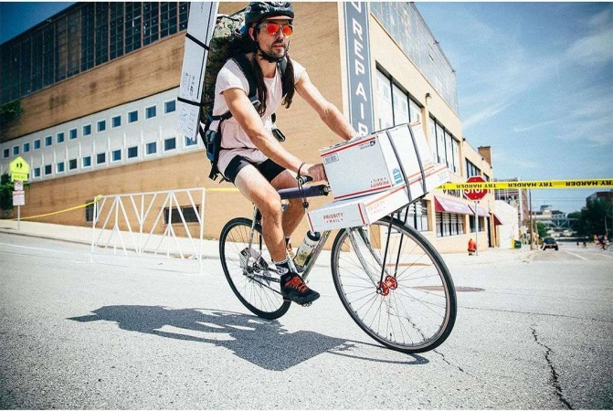 Adult male on a bicycle with boxes attached to the front, wearing helmet and backpack, riding on a city street with buildings.