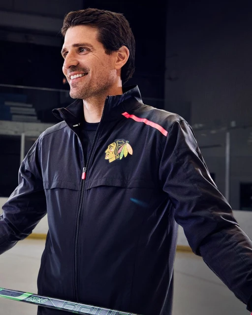Adult man in a Chicago Blackhawks jacket and athletic wear, stands indoors holding a hockey stick and smiling, medium shot.