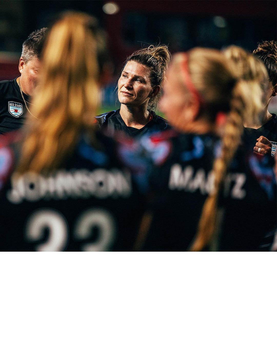 Female soccer player stands with team members, looking hopeful, wearing a black and blue jersey on the sports field.
