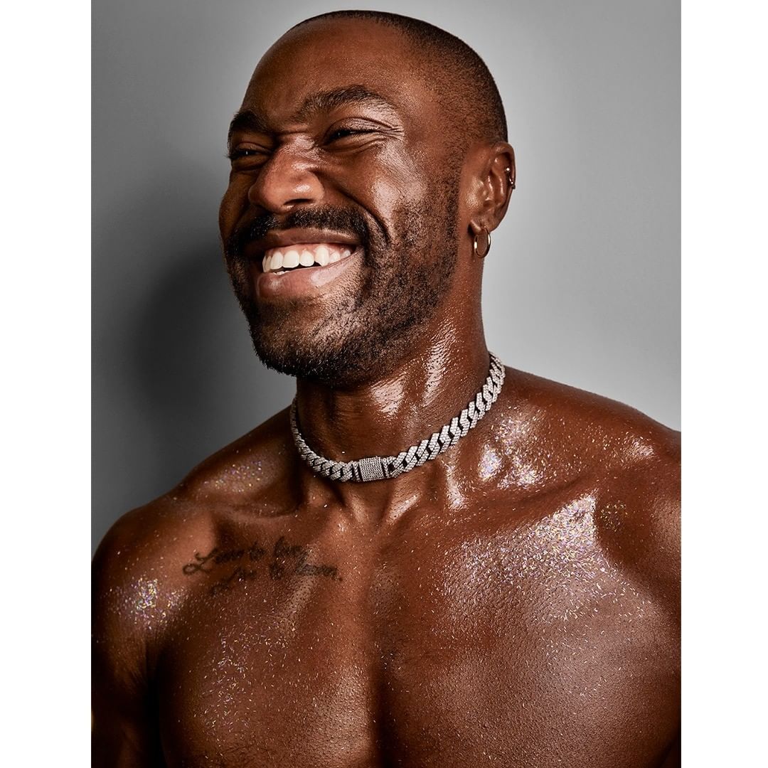 Close-up of a smiling shirtless African-American man with a shaved head and a diamond choker against a gray background.