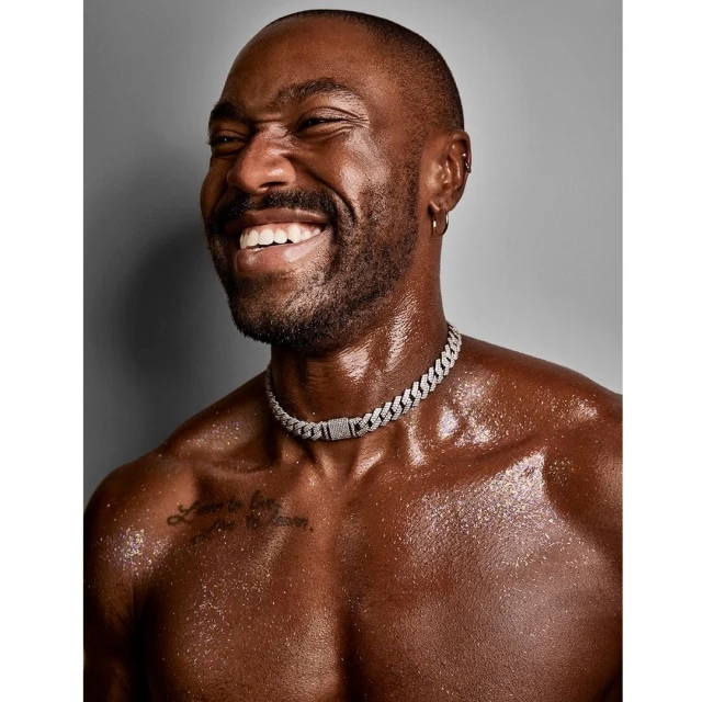 Close-up of a smiling shirtless African-American man with a shaved head and a diamond choker against a gray background.
