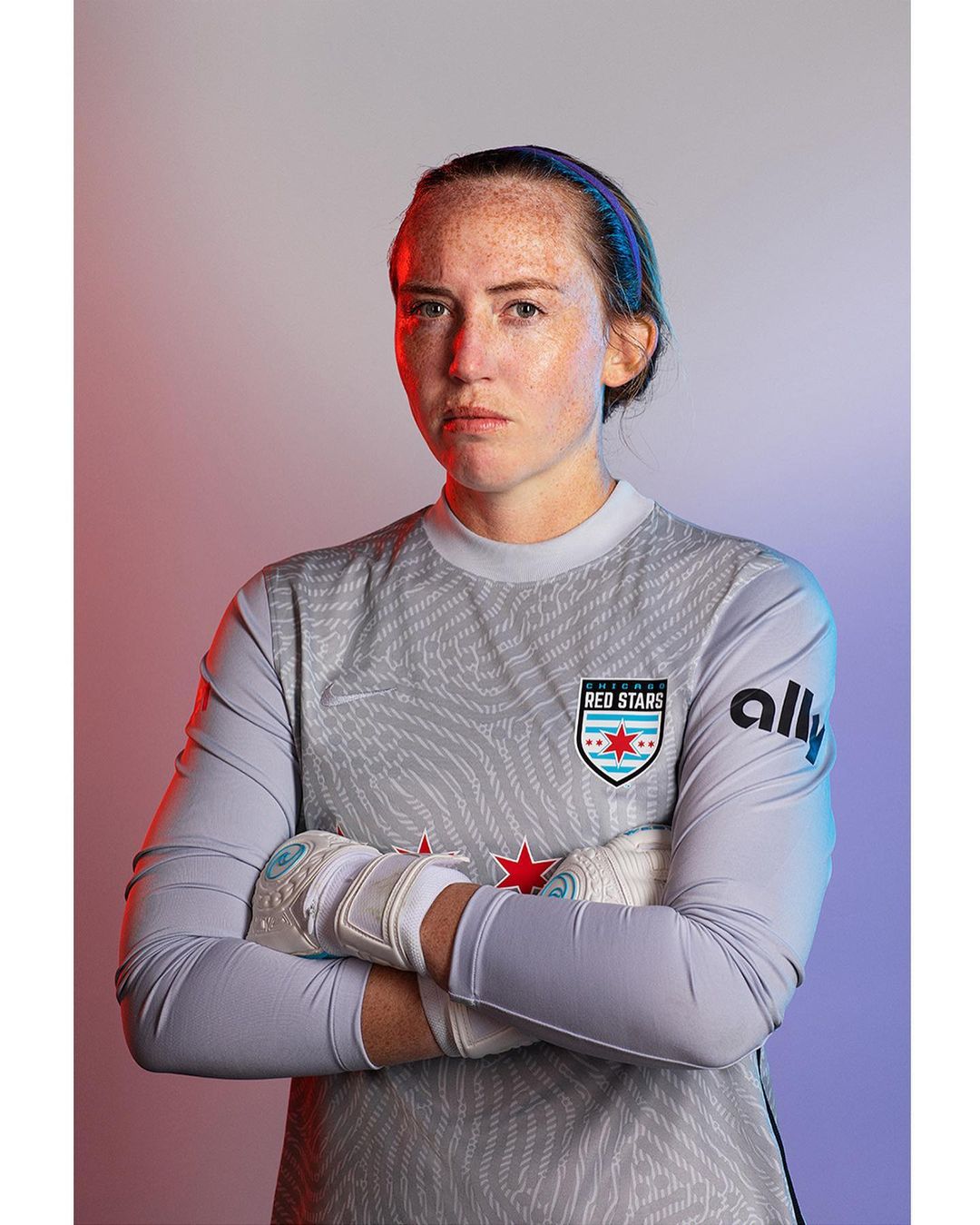 Headshot of a serious adult female athlete in a grey jersey with white goalie gloves with red and blue studio lighting. 