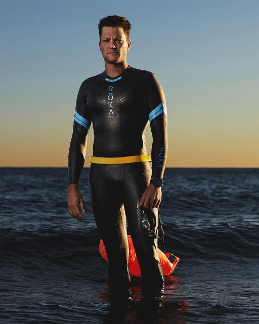 A male athlete in a wetsuit stands waist-deep in ocean water at dusk with a swimming buoy, facing forward in a medium shot.