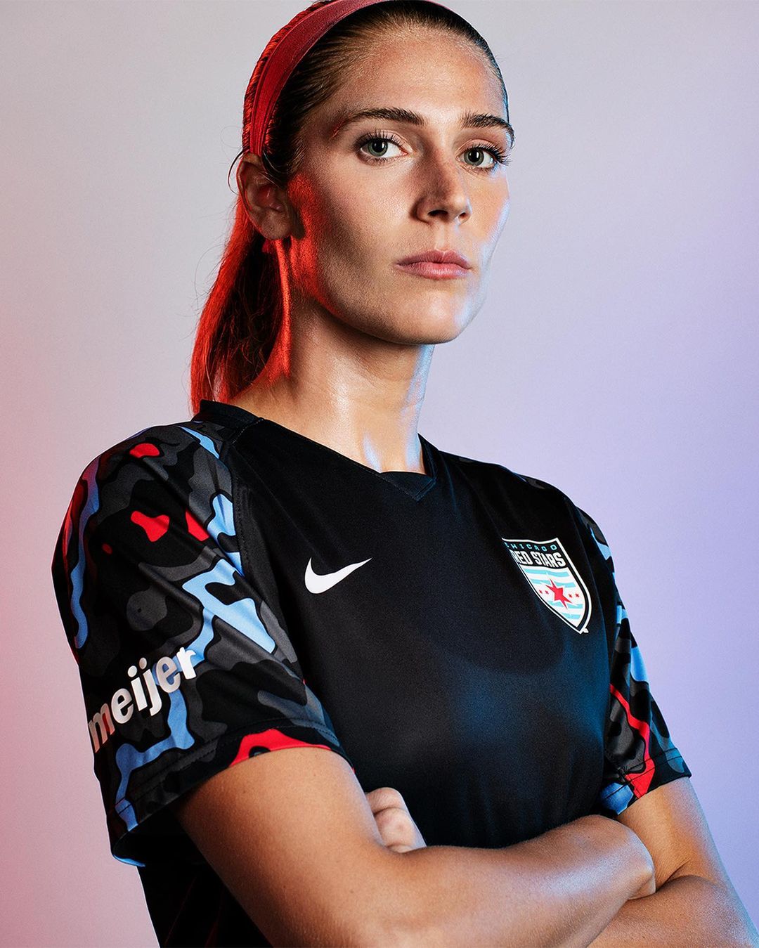 Headshot of a woman in a soccer jersey with arms crossed, set against a gradient background with soft, diffused lighting.