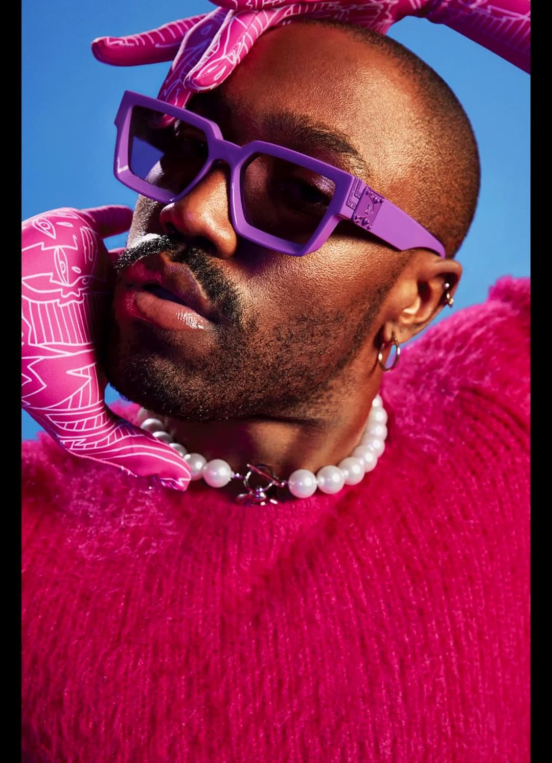 Portrait of adult male in magenta sweater, purple sunglasses and beaded necklace, with magenta patterned gloves and blue background.