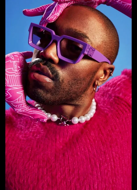 Portrait of adult male in magenta sweater, purple sunglasses and beaded necklace, with magenta patterned gloves and blue background.