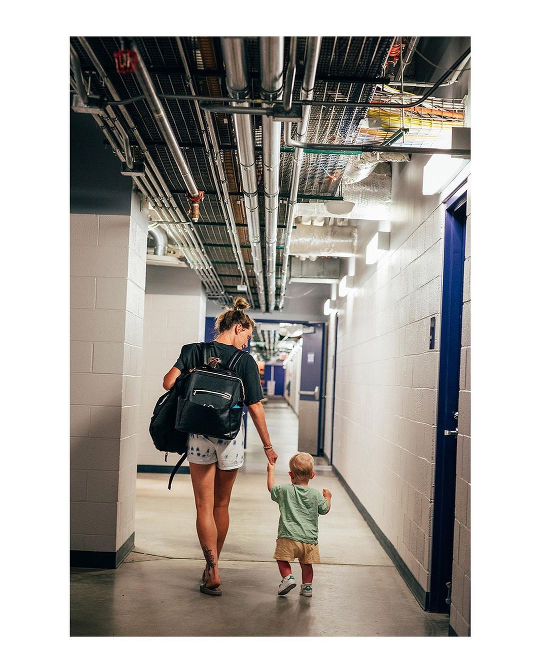 A woman and toddler walk away from the camera down a corridor lined with doors and exposed utility pipes.