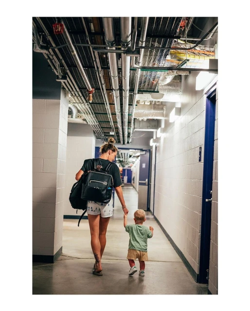 A woman and toddler walk away from the camera down a corridor lined with doors and exposed utility pipes.