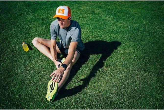 Athlete stretching on green grass, wearing a baseball cap and a grey t-shirt, bottle of energy drink, sunny, outdoor, fitness.