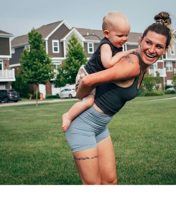 Smiling adult female carrying a toddler on her back in a grassy suburban neighborhood on a sunny day with houses visible.