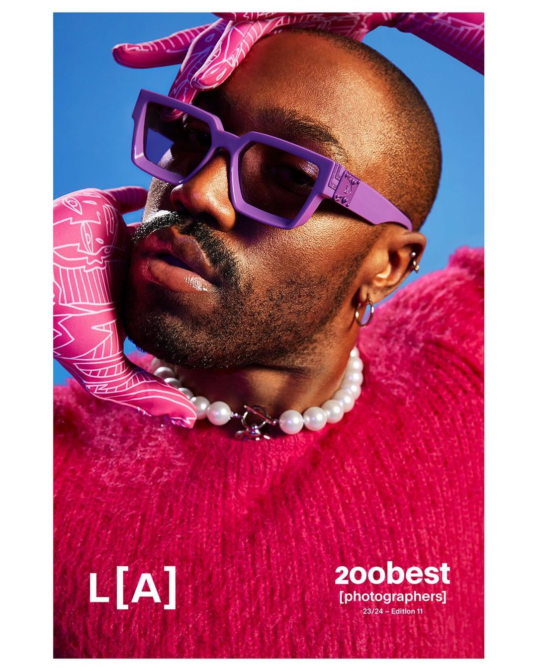 Stylish studio portrait of an African-American male model wearing lavender sunglasses and a pearl necklace against a vibrant blue backdrop.