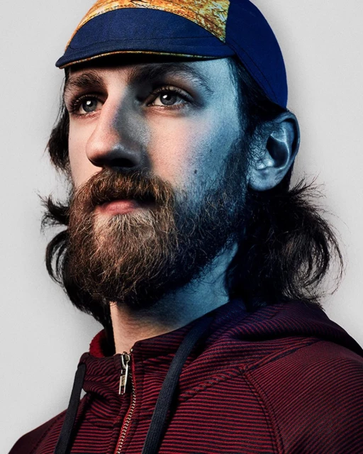 Close-up portrait of a bearded adult male with a blue and gold cycling cap, wearing a maroon hooded sweatshirt in a studio setting.