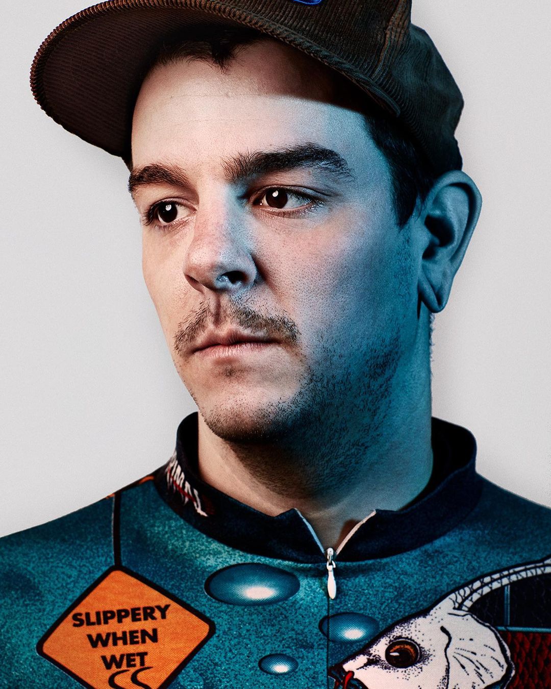 Portrait of a young adult male wearing a brown corduroy cap and printed top against a white background in eye-level, medium shot.