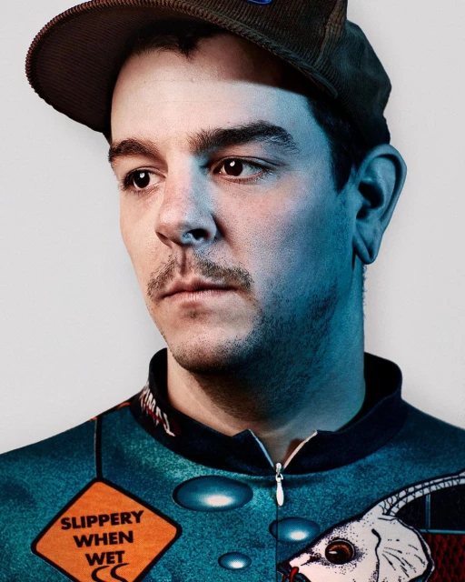 Portrait of a young adult male wearing a brown corduroy cap and printed top against a white background in eye-level, medium shot.