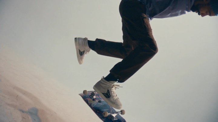Skateboarder in motion mid-air, wearing white shoes and brown pants, caught in a dynamic low-angle shot against a neutral background.