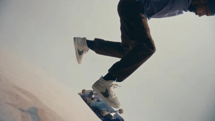 Skateboarder in motion mid-air, wearing white shoes and brown pants, caught in a dynamic low-angle shot against a neutral background.