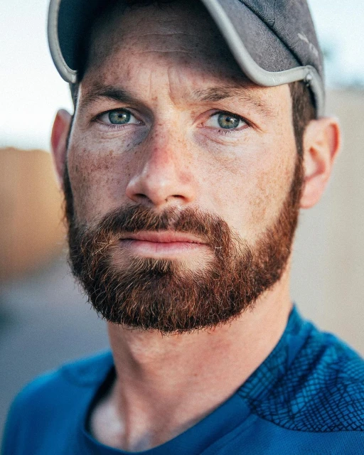 Close-up portrait of a bearded adult male with freckles, wearing a blue shirt and gray baseball cap, captured at eye-level.