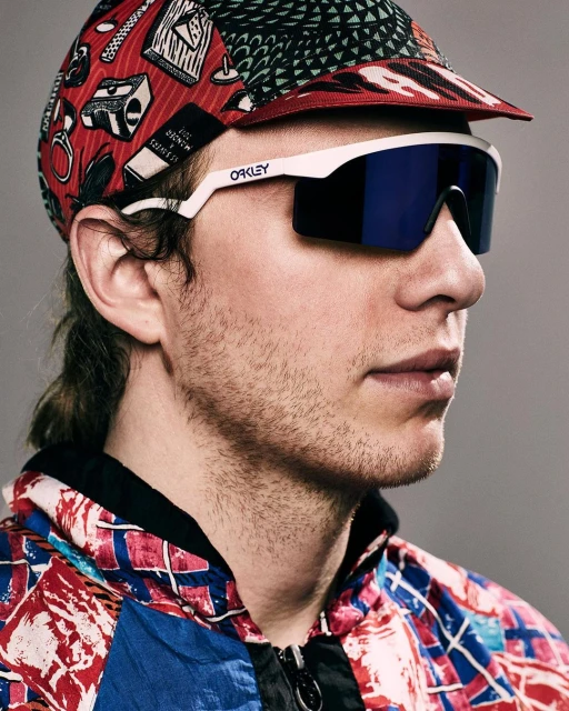 Adult male wearing Oakley sunglasses and patterned cap and jacket against a gray background in medium studio portrait.