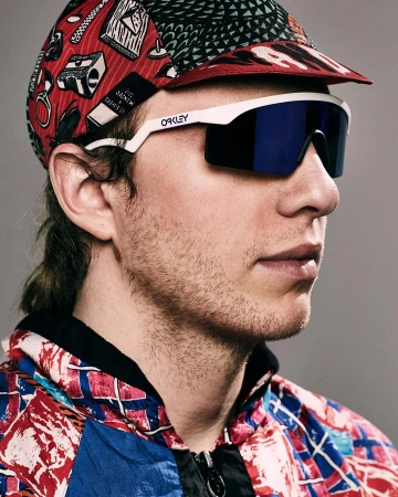 Adult male wearing Oakley sunglasses and patterned cap and jacket against a gray background in medium studio portrait.