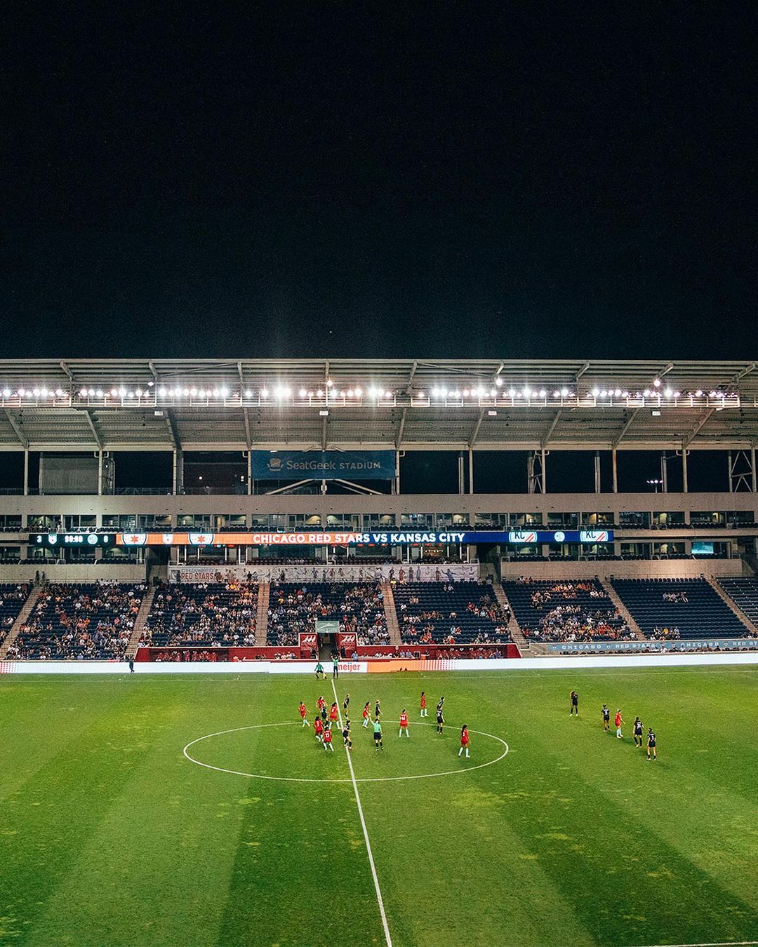Wide shot of a soccer field at night with two teams lined up on the field during a match, stadium lights illuminate the scene.