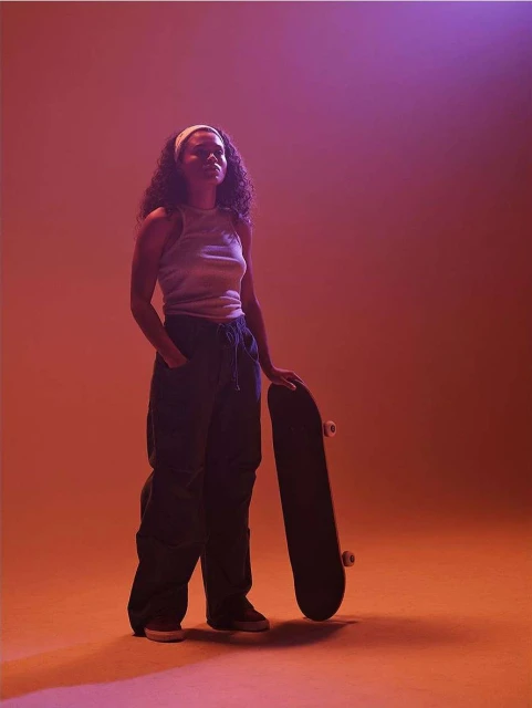 Young woman with a skateboard in a studio with orange and purple lighting, wearing a headband, top, and loose black pants and sneakers.