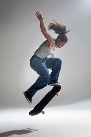 Woman in grey tank top and blue jeans performing a skateboard trick mid-air in front of a white seamless studio background.