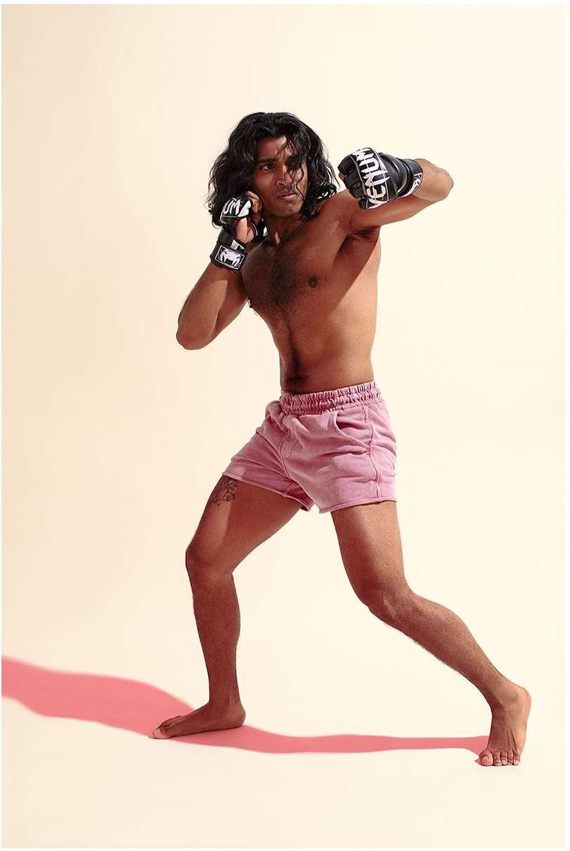 An adult male boxer in pink shorts wearing boxing gloves poses in a fighting stance in a studio with a cream-colored background.