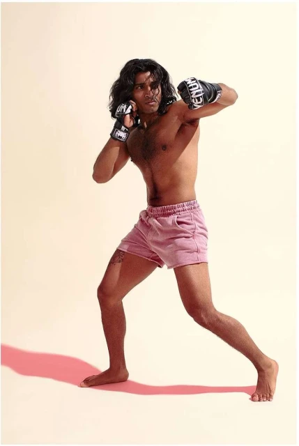 An adult male boxer in pink shorts wearing boxing gloves poses in a fighting stance in a studio with a cream-colored background.