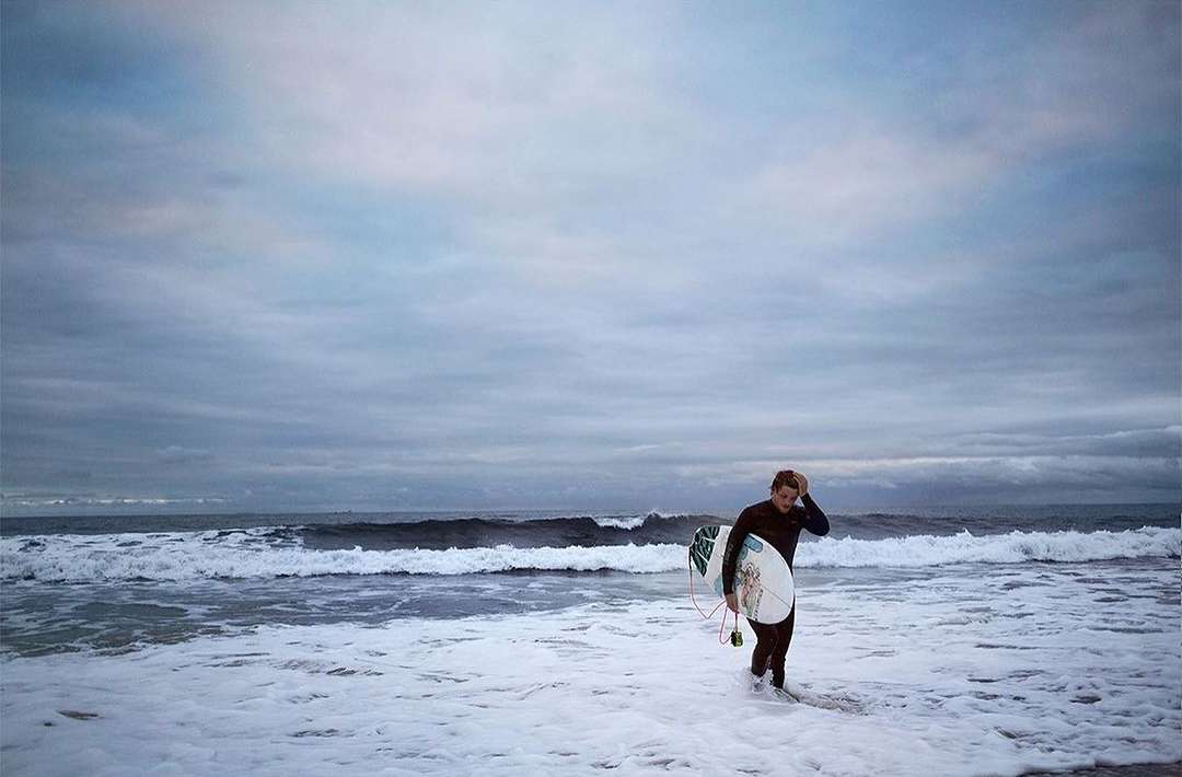 Man in wetsuit walks along beach with surfboard, waves and overcast sky visible. Beach scene with surfer. Sport and outdoor recreation.