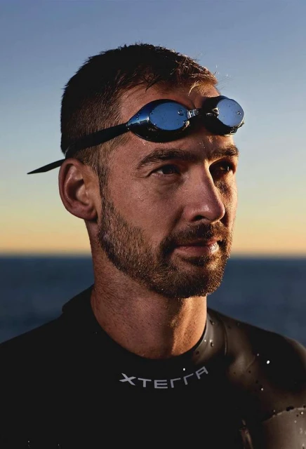 Close-up of a man in a wetsuit and swimming goggles, standing in front of the ocean and sky. He has a beard and is looking to the side.