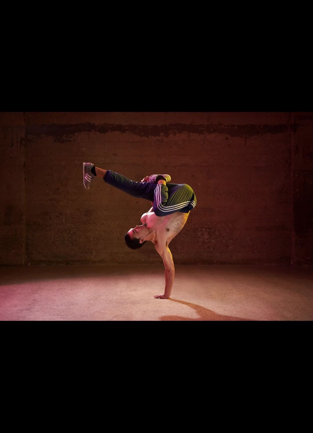 A male dancer balances upside down on one hand in a warehouse-style room. He wears athletic pants and sneakers in this fitness shot.